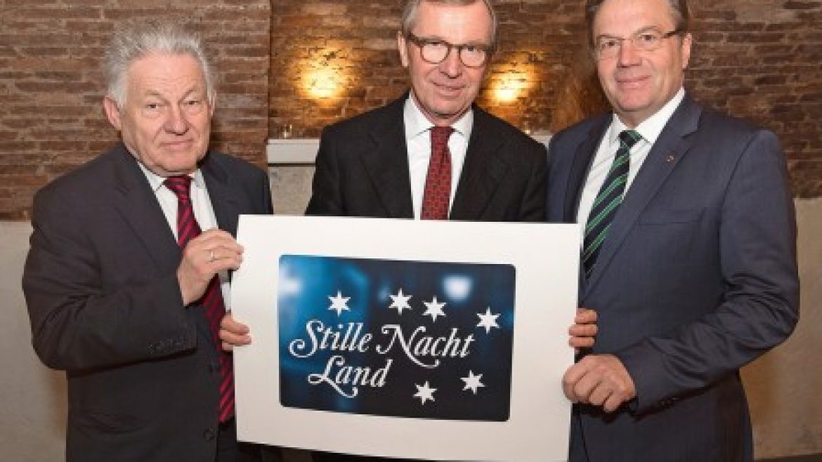 Drei Männer in Anzügen halten ein Schild mit der Aufschrift "Stille Nacht Land" mit Sternen darauf und stehen in einem Haus.