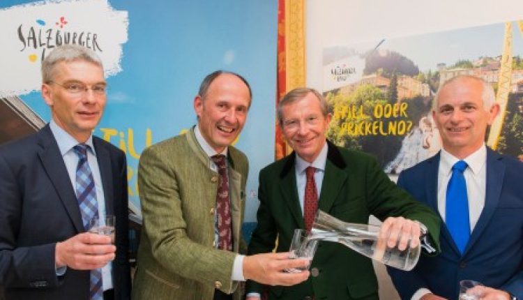 Vier Männer in Anzügen halten oder schenken Gläser mit Wasser ein und lächeln bei einer Werbeveranstaltung in einer Halle.