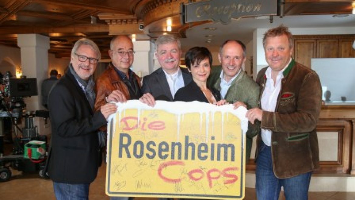 In einem hölzernen Empfangsraum halten sechs Personen ein Die Rosenheim Cops-Schild und lächeln in die Kamera.