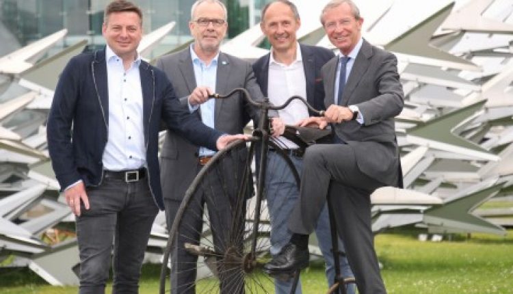 Vier Männer in Anzügen lächeln und posieren mit einem altmodischen Fahrrad vor einem modernen Gebäude und einer Skulptur.