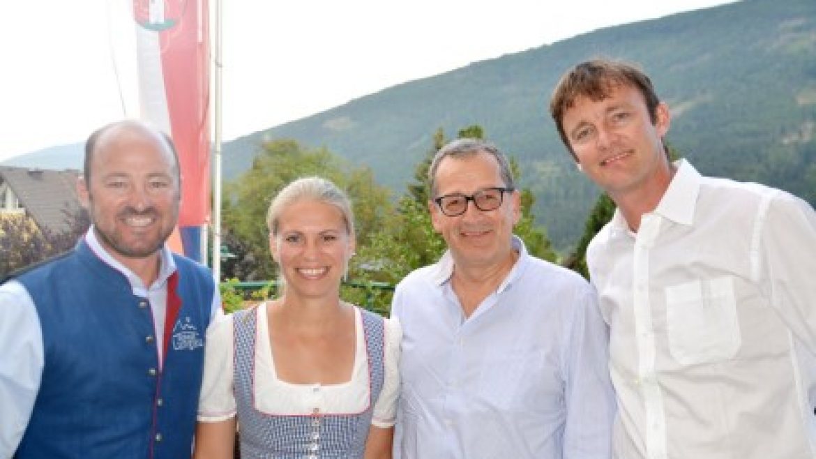 Vier lächelnde Erwachsene posieren im Freien mit Bergen im Hintergrund; eine Frau trägt ein traditionelles österreichisches Kleid.