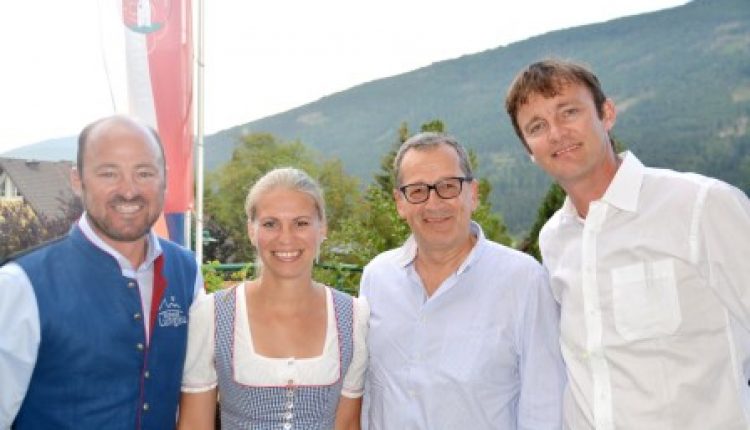 Vier lächelnde Erwachsene posieren im Freien mit Bergen im Hintergrund; eine Frau trägt ein traditionelles österreichisches Kleid.