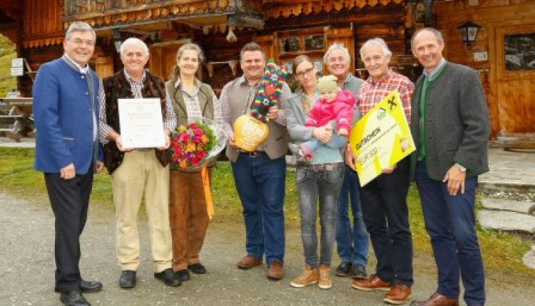 Eine Gruppe von Menschen steht lächelnd vor einem rustikalen Holzgebäude und hält Blumen und Urkunden in der Hand.