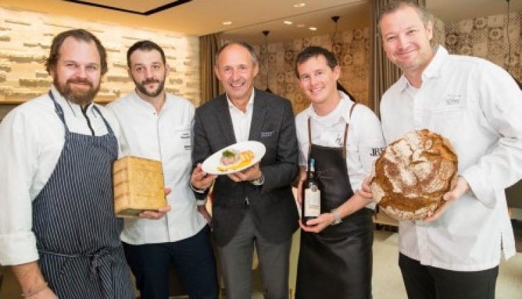 Fünf lächelnde Männer, die Brot, einen Teller und eine Flasche in der Hand halten, stehen in einem Restaurant zusammen.