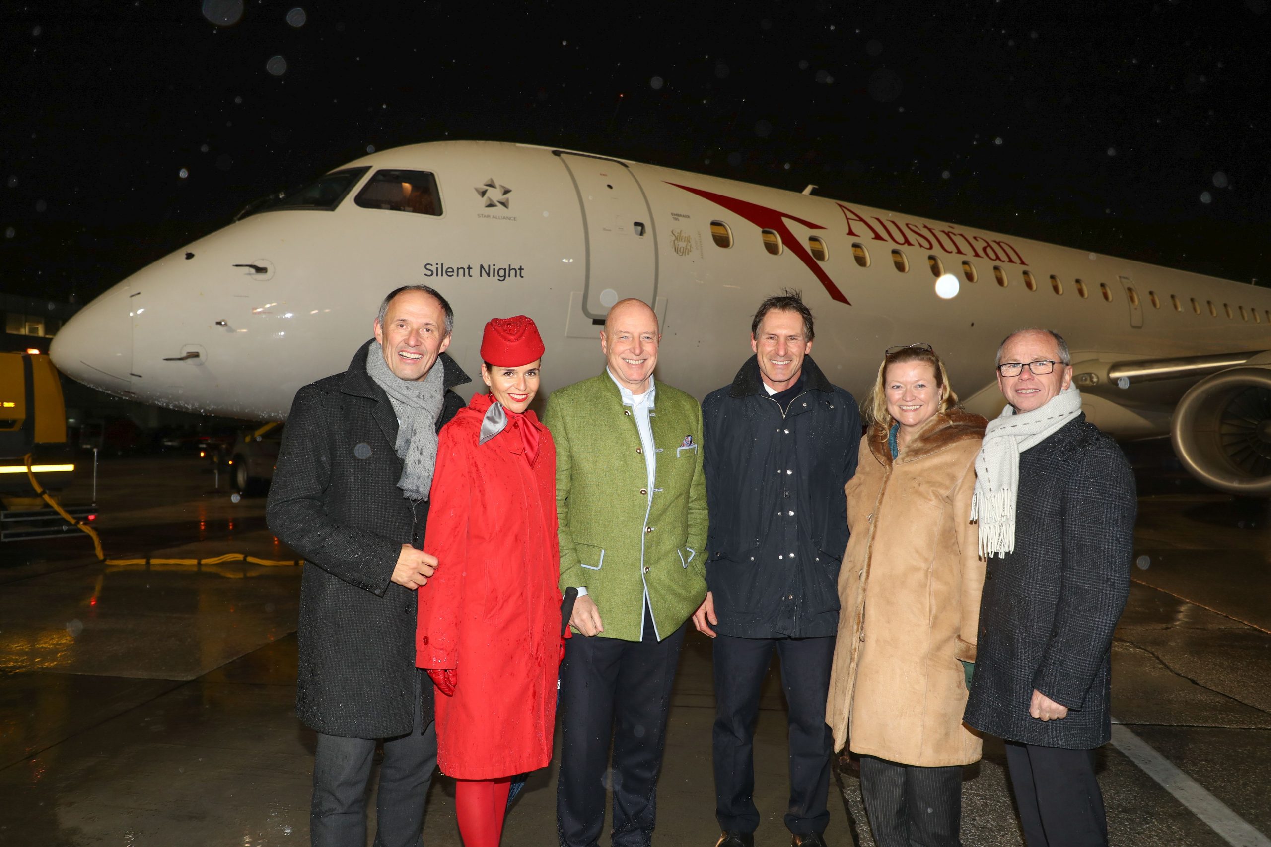 Sechs Personen posieren lächelnd vor einem Flugzeug der Austrian Airlines, das nachts auf einer nassen Landebahn steht.