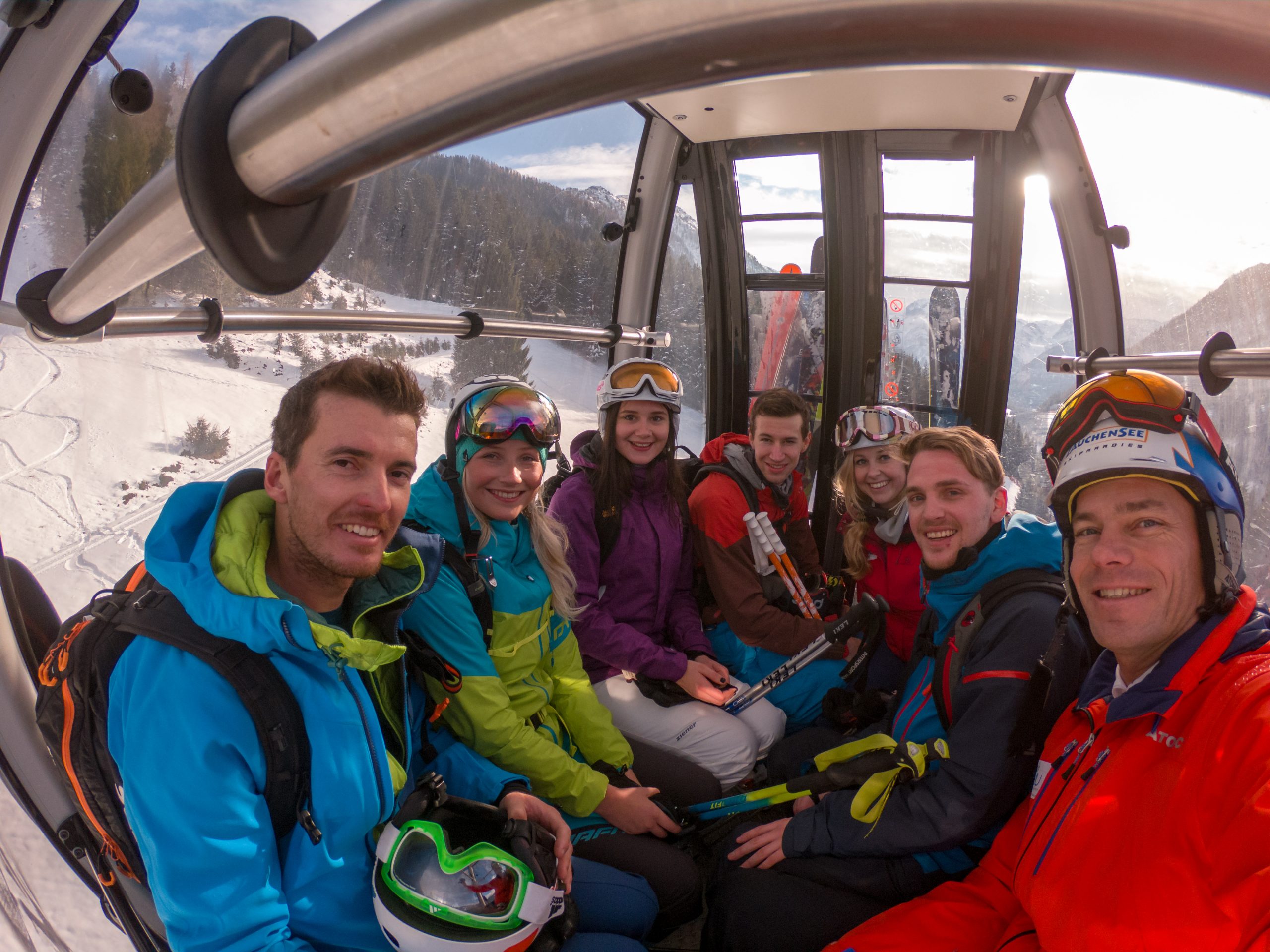 Sieben lächelnde Skifahrer in farbenfroher Kleidung fahren in einer Skigondel, durch deren Fenster verschneite Berge zu sehen sind.