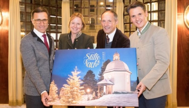 Vier Personen, die lächelnd ein Stille-Nacht-Gemälde einer verschneiten Kapelle mit Weihnachtsbäumen halten.