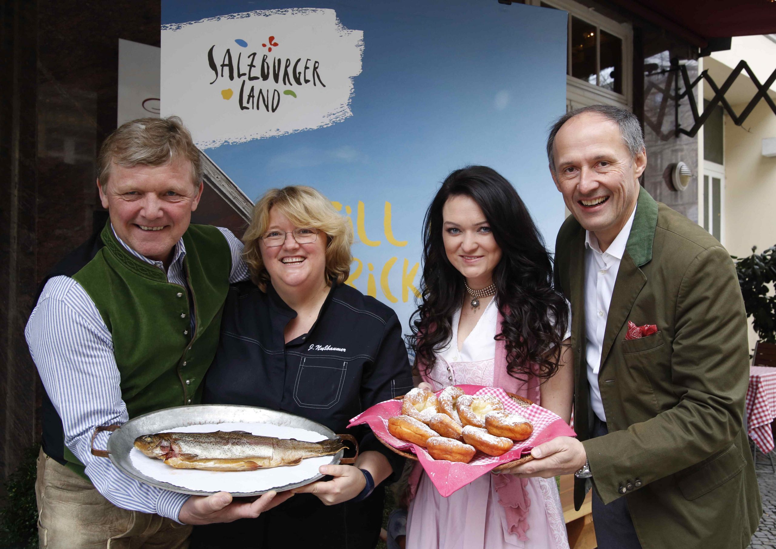Vier lächelnde Menschen mit Tellern voller Fisch und Gebäck vor einem Salzburger Tourismusbanner.