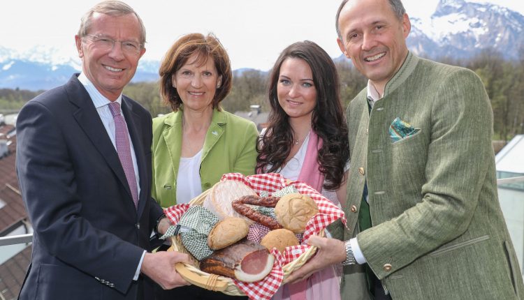 Vier lächelnde Menschen in österreichischer Tracht halten einen Korb mit Brot und Würstchen, im Hintergrund sind die Berge zu sehen.