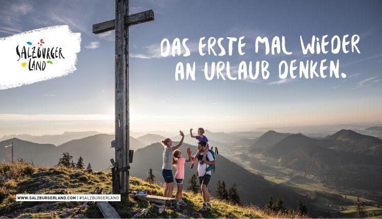 Eine Familie steht bei Sonnenuntergang vor einem Bergkreuz und gibt sich die Hand. Text: Das erste Mal wieder an Urlaub denken.