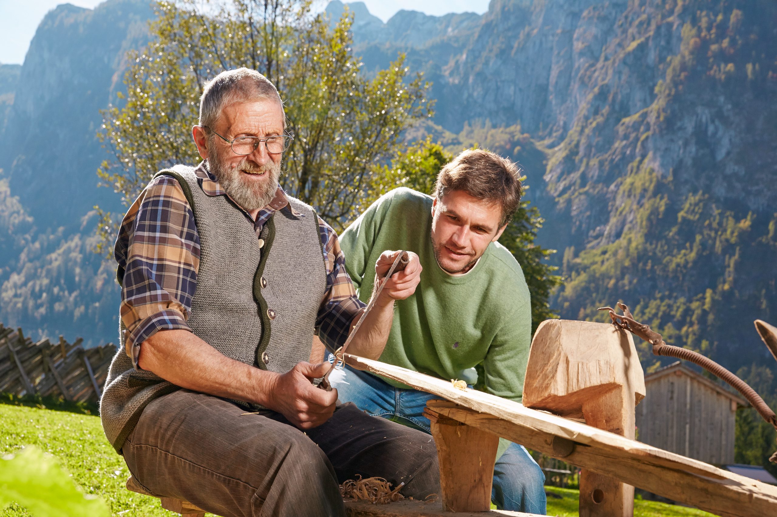 Ein älterer Mann schnitzt Holz beim Bauernherbst, während ein jüngerer Mann zusieht, mit den Bergen im Hintergrund.