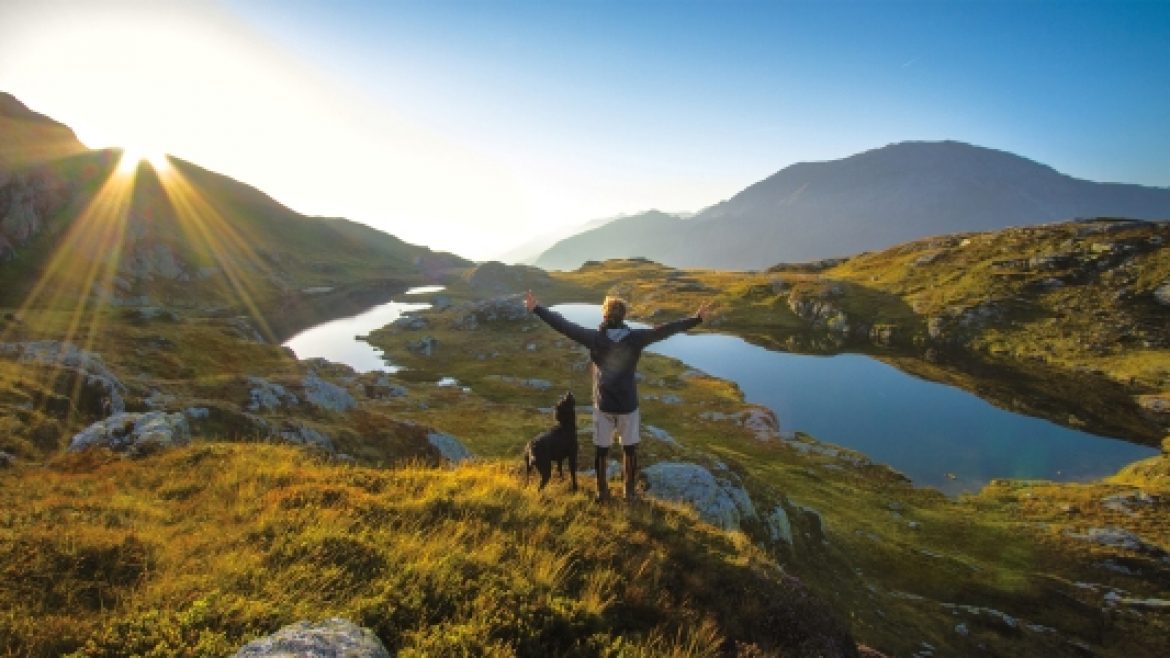 Person mit ausgestreckten Armen und einem Hund auf einem Hügel, die den Sonnenaufgang und die Naturtrends über Seen und Bergen genießen.
