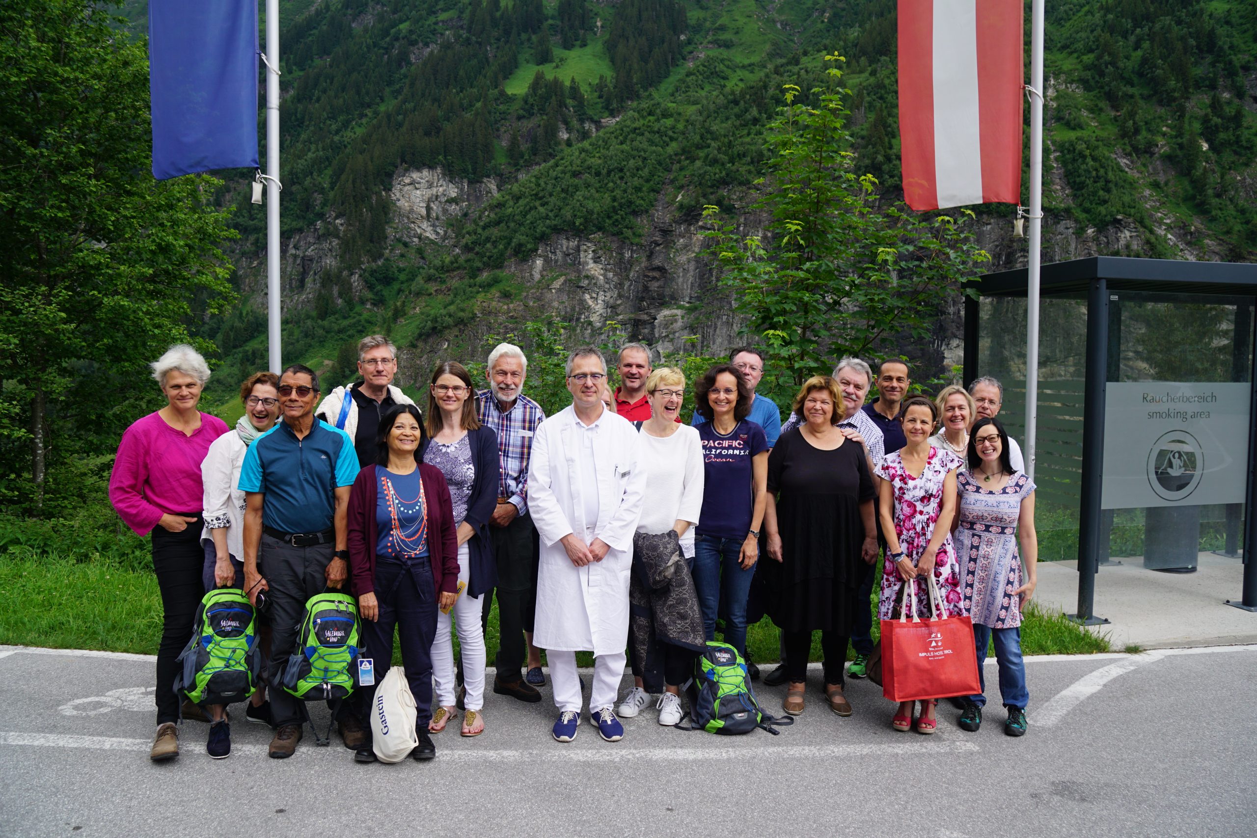 Eine Gruppe von Menschen auf einer Ärztestudienreise posiert im Freien in der Nähe von Fahnen und Bergen, im Hintergrund ist Grün zu sehen.
