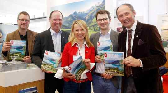 Fünf Menschen lächeln und halten Reisemagazine vor einer malerischen Schweizer Landschaft.