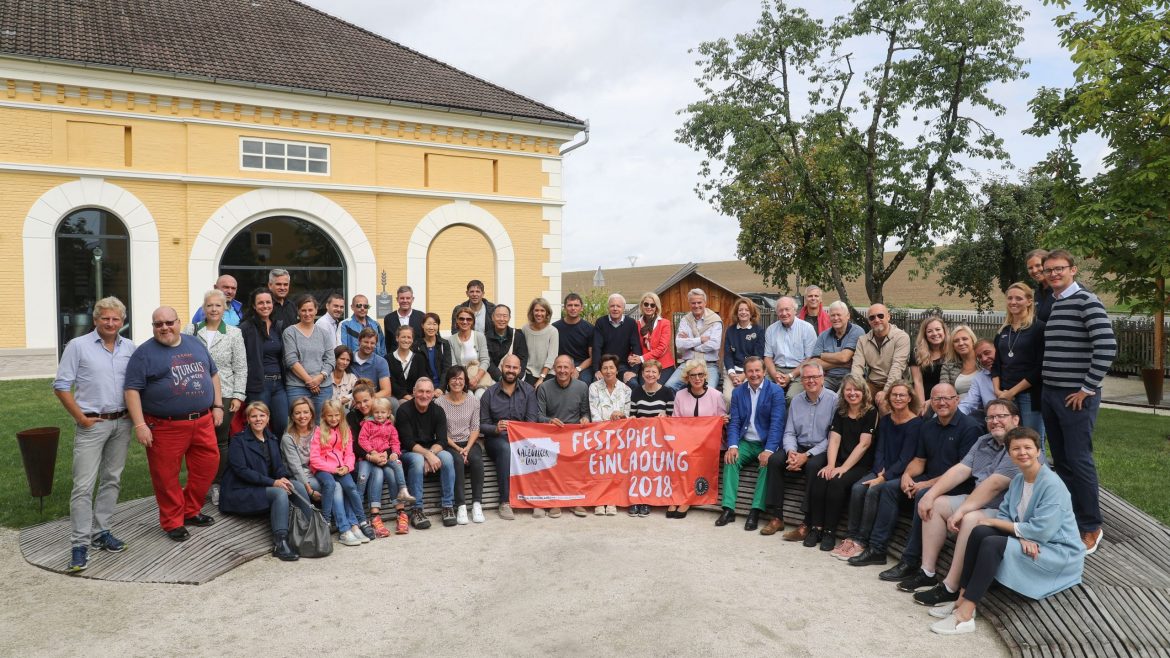 Eine große Gruppe lächelnder Menschen posiert im Freien und hält ein rotes Banner mit der Aufschrift "Festspiel Einladung 2018".