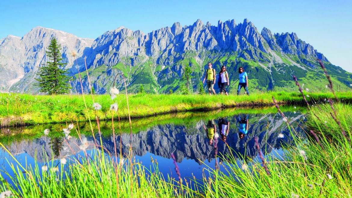 Vier Personen wandern an einem Teich, in dem sich die Berge und der blaue Himmel spiegeln.