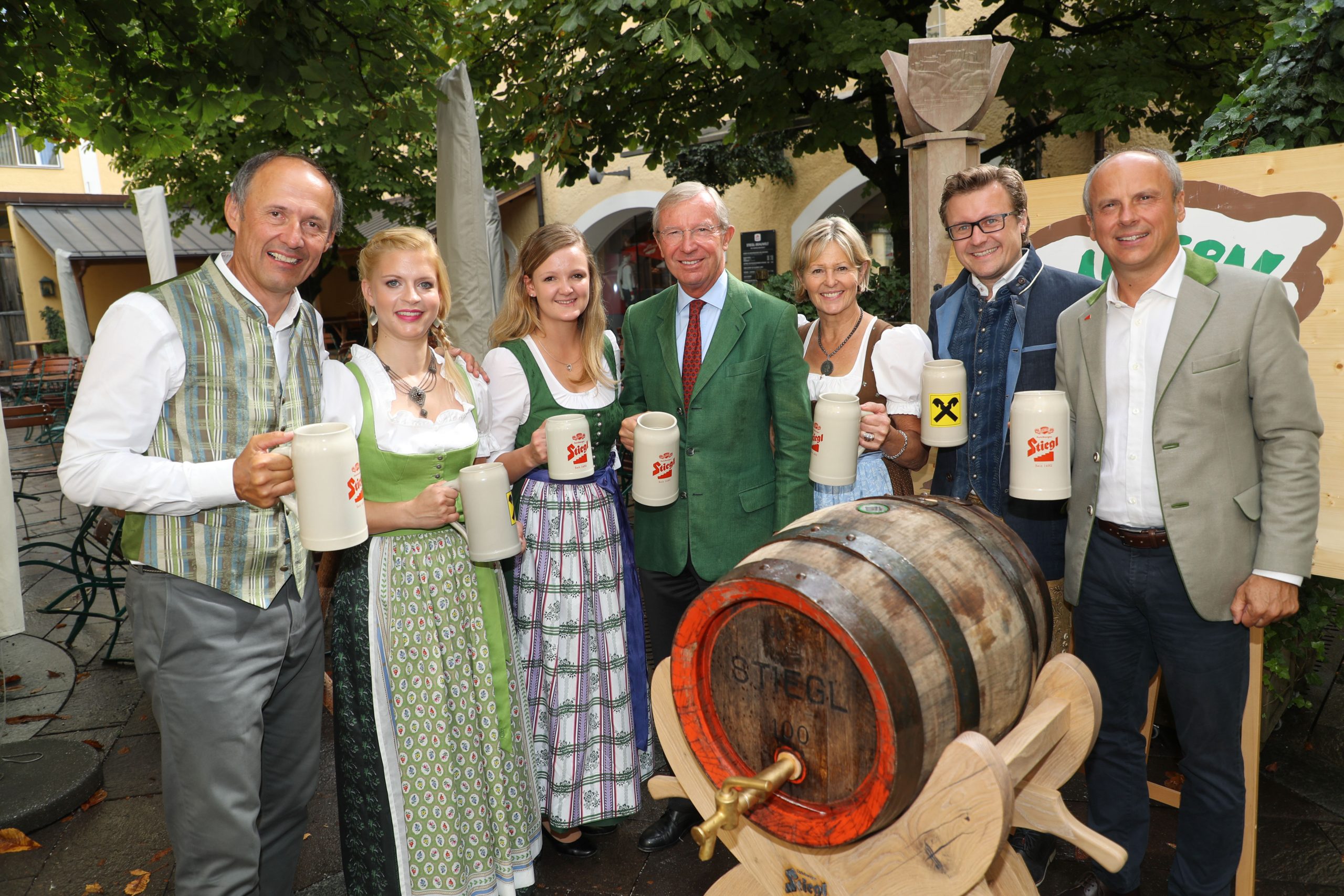 Sieben Personen in traditioneller österreichischer Kleidung halten Bierkrüge und stehen an einem hölzernen Bierfass im Freien.