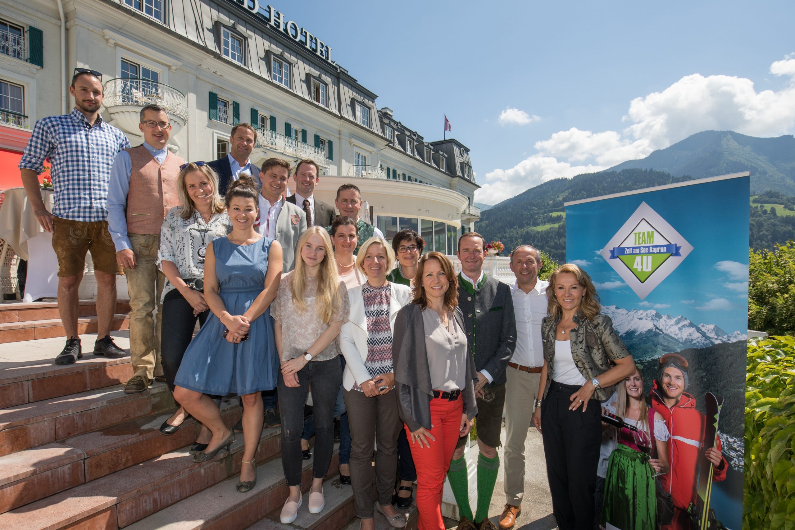 Eine Gruppe von Menschen posiert an einem sonnigen Tag gemeinsam vor einem Hotel mit Bergen im Hintergrund.