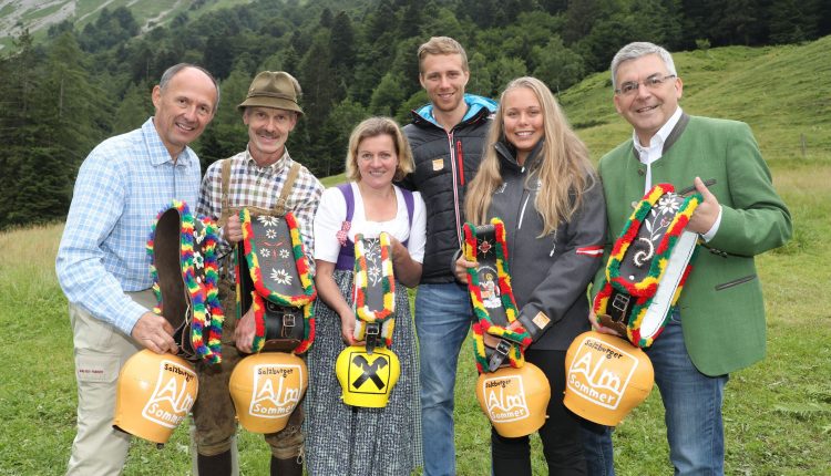 Sechs Menschen in traditioneller Kleidung halten bunte Kuhglocken und lächeln in die Kamera, im Hintergrund grüne Hügel.