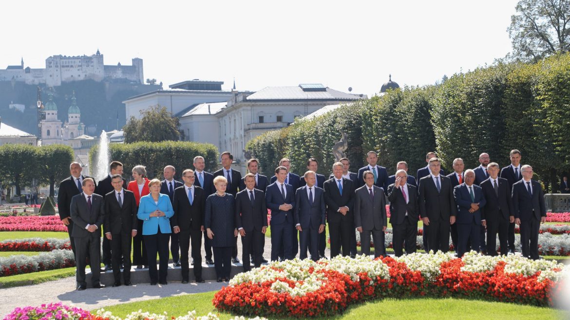 Eine Gruppe von Personen in formeller Kleidung posiert für ein Foto in einem Garten mit Blumen und historischen Gebäuden im Hintergrund.