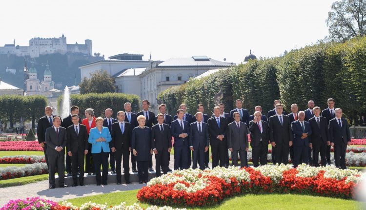 Eine Gruppe von Personen in formeller Kleidung posiert für ein Foto in einem Garten mit Blumen und historischen Gebäuden im Hintergrund.
