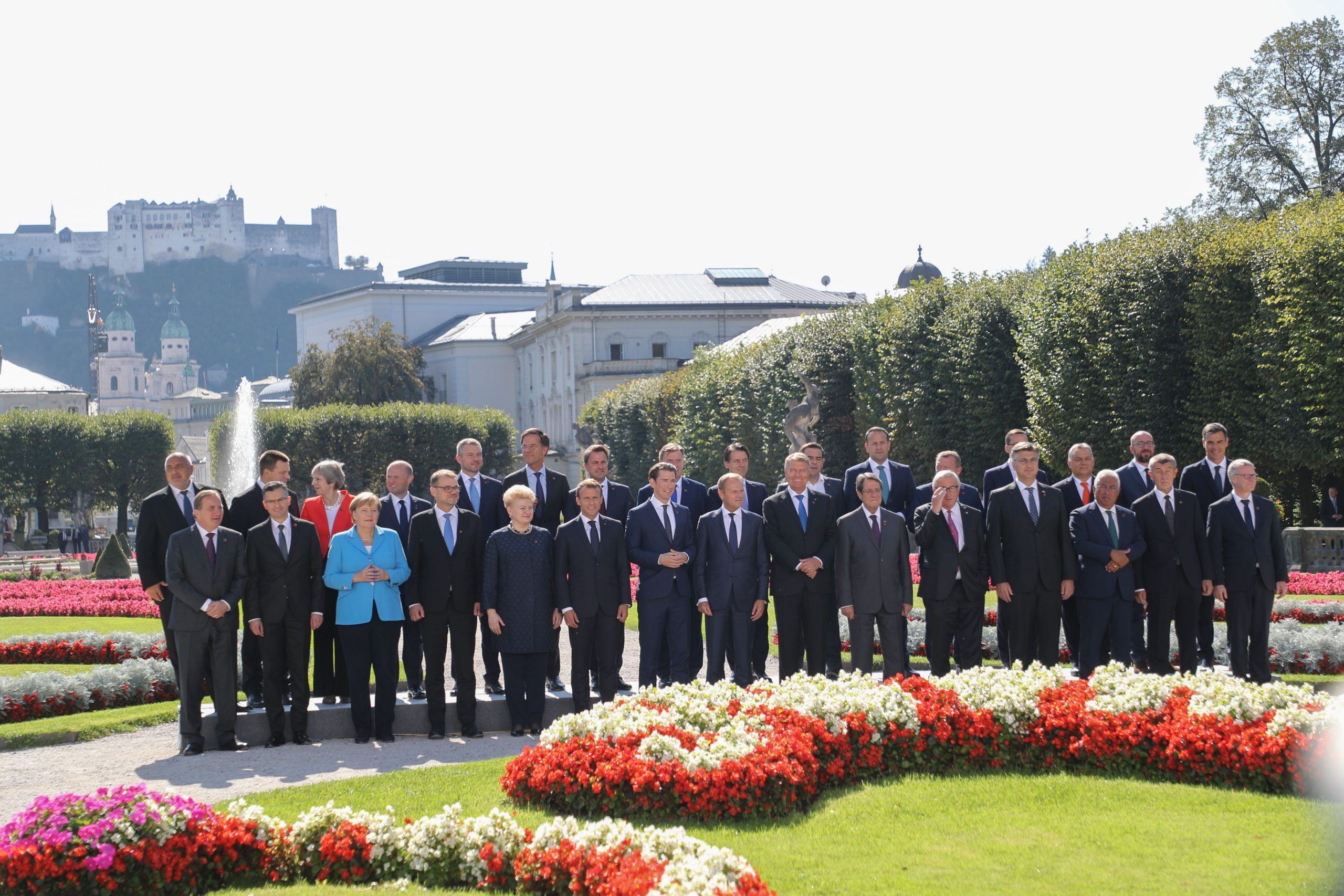 Eine Gruppe von Personen in formeller Kleidung posiert für ein Foto in einem Garten mit Blumen und historischen Gebäuden im Hintergrund.