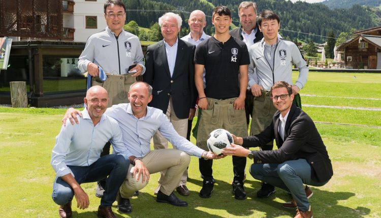 Eine Gruppe von Männern posiert und lächelt im Freien auf einer Wiese, wobei zwei Männer vorne einen Fußball halten.