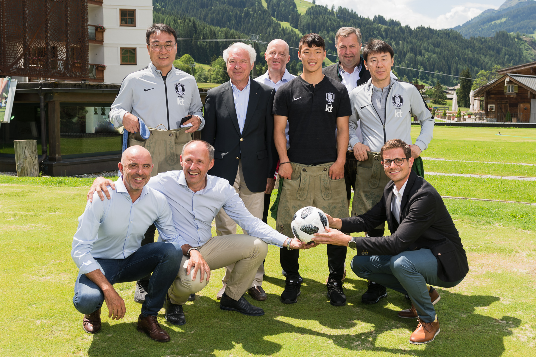 Eine Gruppe von Männern posiert und lächelt im Freien auf einer Wiese, wobei zwei Männer vorne einen Fußball halten.
