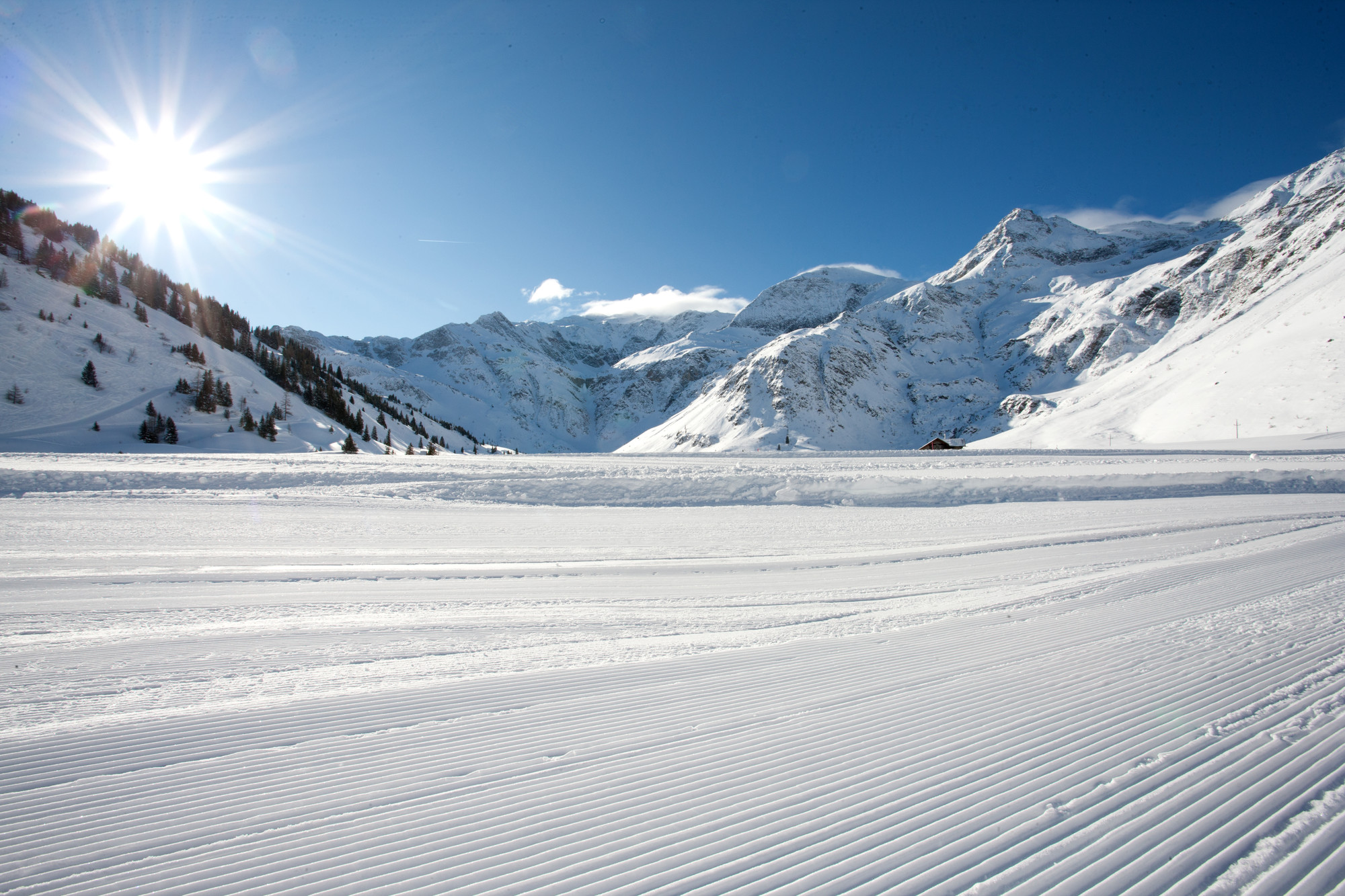 Die Sonne scheint auf eine verschneite Berglandschaft mit präparierten Skipisten im Vordergrund.