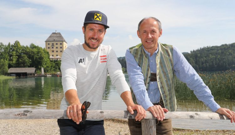 Zwei Männer lächeln an einem See mit einem Gebäude und Bäumen im Hintergrund an einem sonnigen Tag.