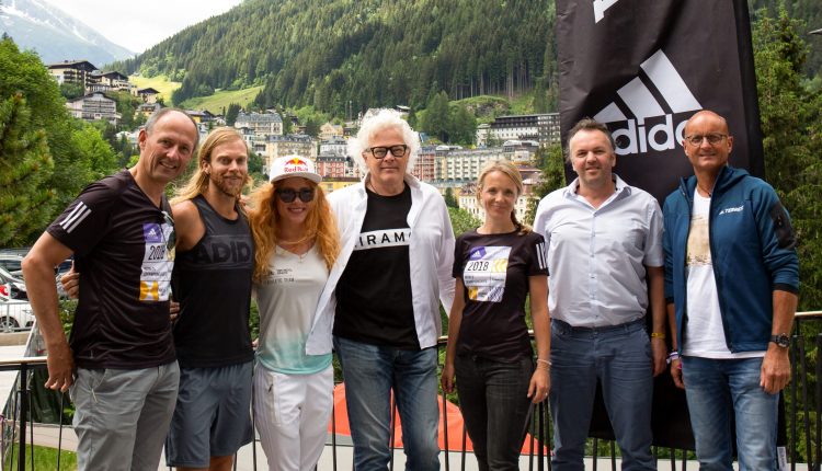 Sieben Personen stehen im Freien vor Bergen und Gebäuden, neben einer großen Adidas-Flagge und lächeln in die Kamera.