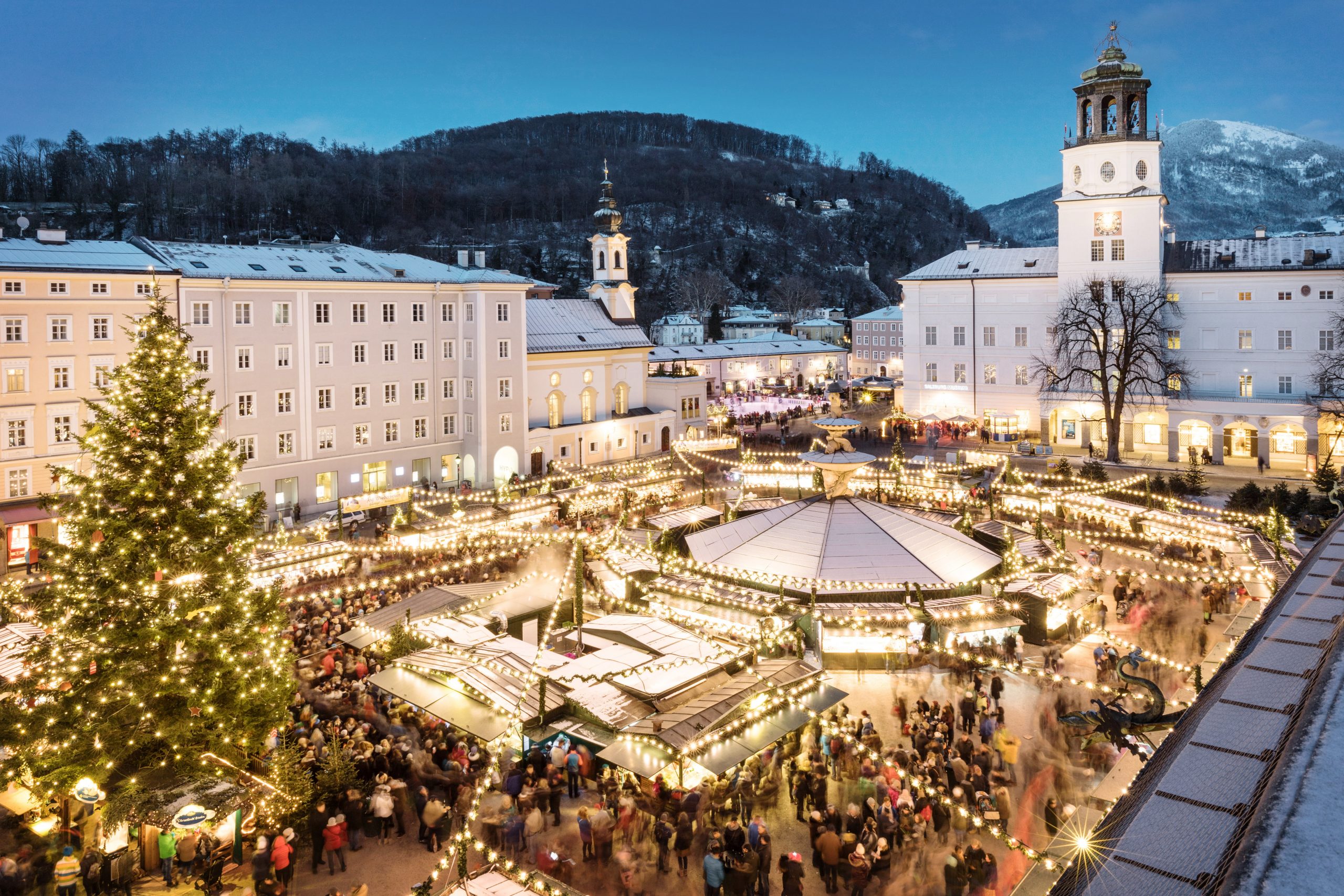 Ein festlicher Weihnachtsmarkt in der Abenddämmerung mit Lichtern, einem großen Baum und vielen Menschen auf einem verschneiten Stadtplatz.