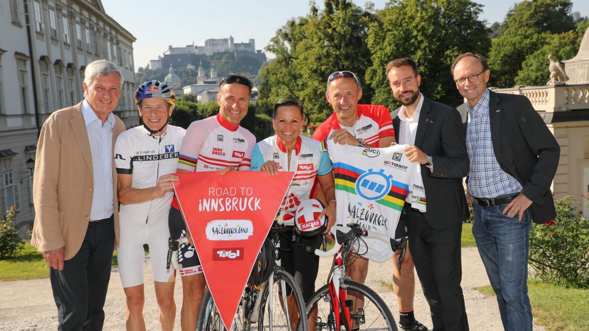 Sieben lächelnde Menschen, einige in Radfahrerkleidung, posieren mit Fahrrädern und einem roten Road to Innsbruck-Banner im Freien.