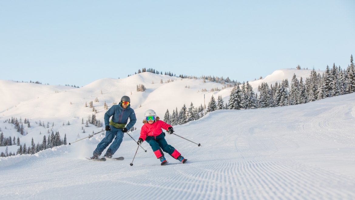 Ein Erwachsener und ein Kind fahren auf einer präparierten, verschneiten Piste mit Kiefern und Bergen im Hintergrund.