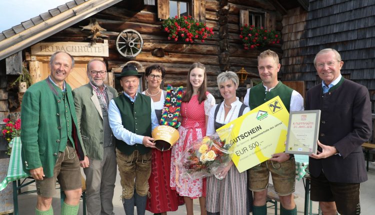Acht Personen in traditioneller österreichischer Tracht stehen lächelnd mit Geschenken vor einem rustikalen Holzgebäude.