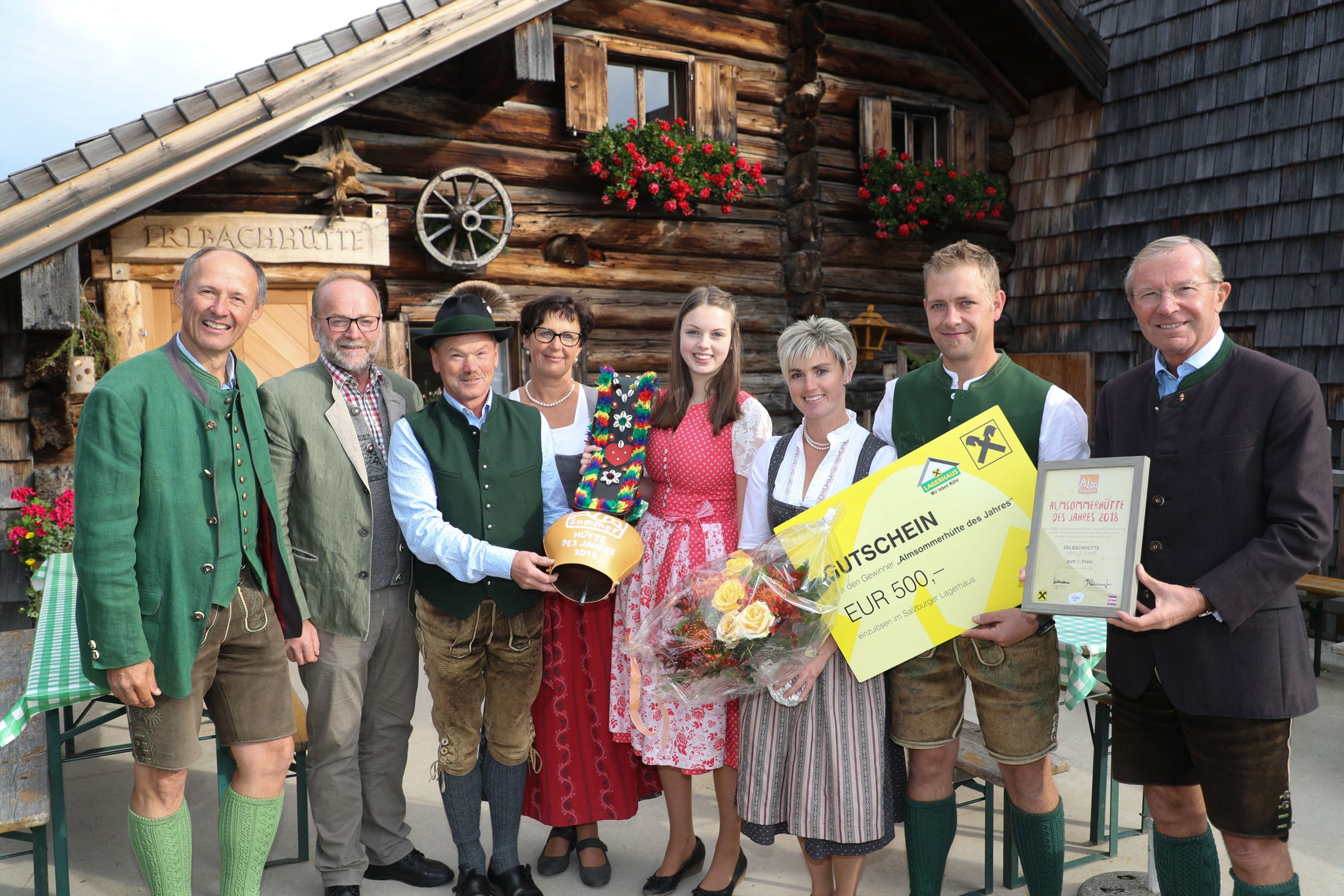 Acht Personen in traditioneller österreichischer Tracht stehen lächelnd mit Geschenken vor einem rustikalen Holzgebäude.