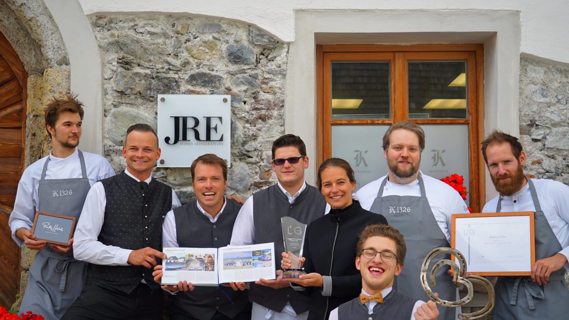 Ein lächelndes Restaurantteam posiert mit Auszeichnungen und Urkunden vor einem Steingebäude mit einem JRE-Schild.