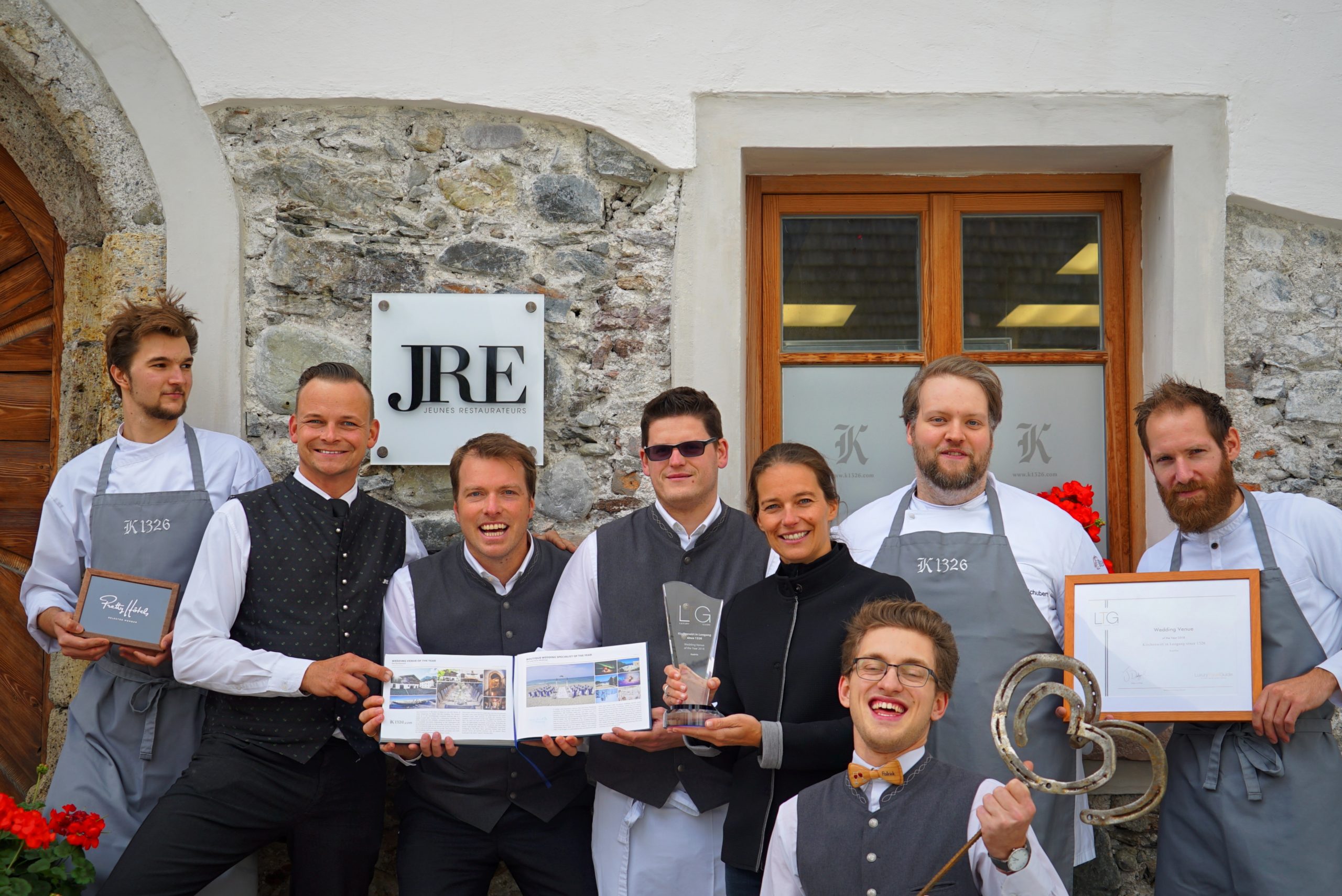Ein lächelndes Restaurantteam posiert mit Auszeichnungen und Urkunden vor einem Steingebäude mit einem JRE-Schild.