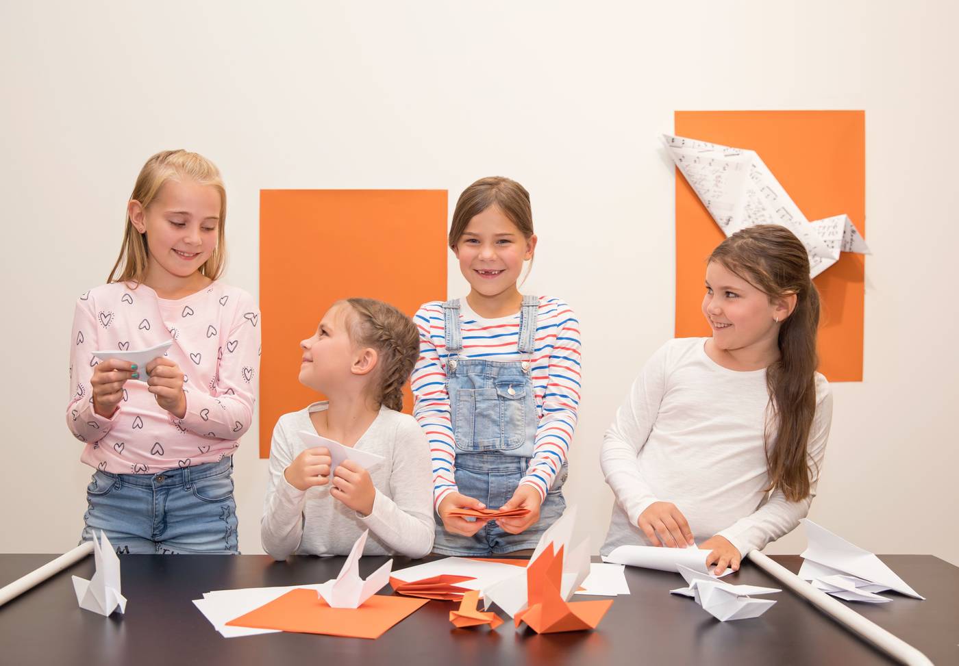 Vier junge Mädchen lächeln, während sie an einem Tisch mit orangefarbenem Papier und Origami-Kunstwerken an der Wand hinter ihnen Papier-Origami basteln.