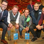 Fünf Männer posieren in einem Innenraum, kniend und lächelnd, mit kleinen blauen Fahnen vor ihnen.