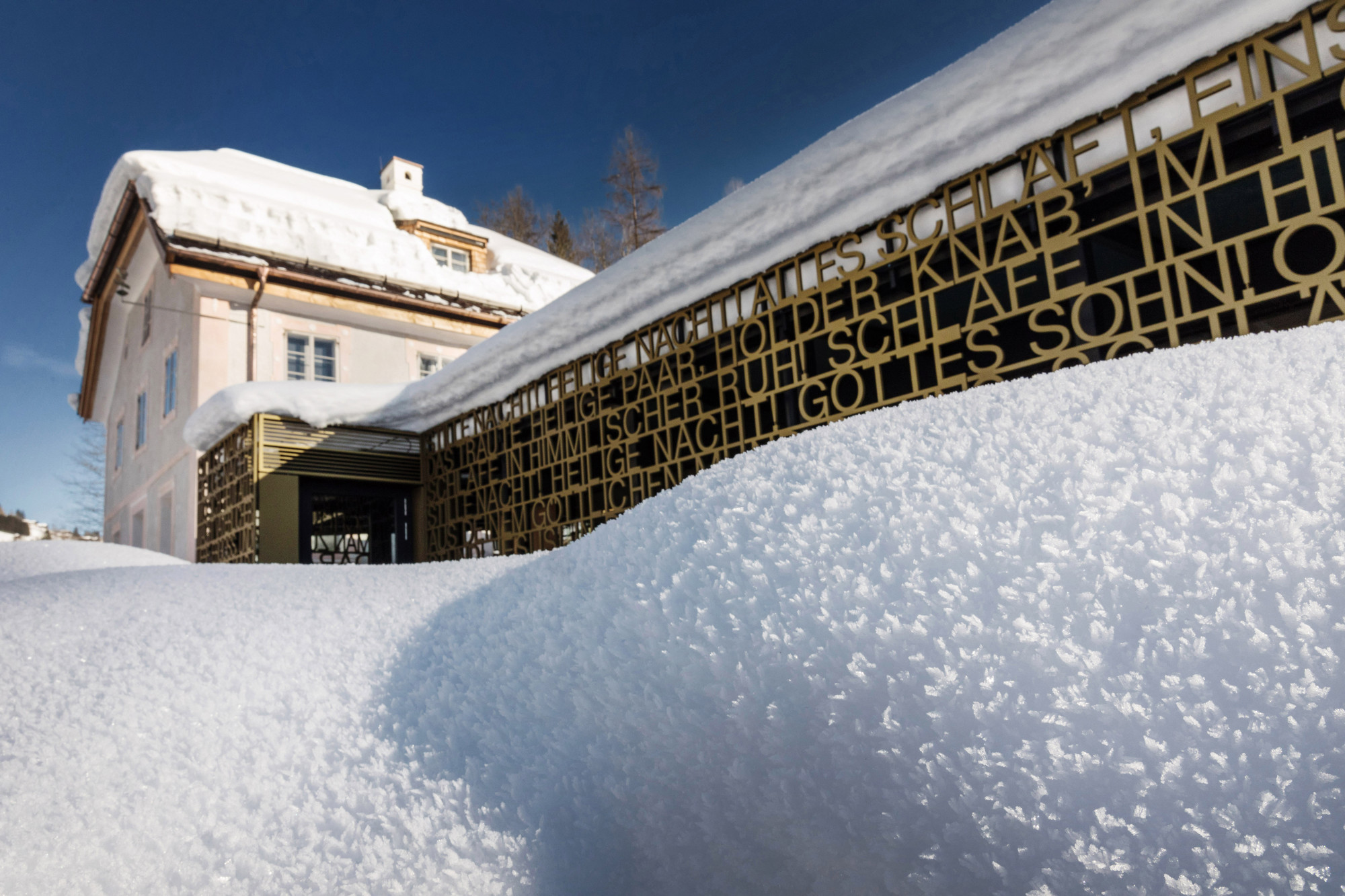 Ein schneebedecktes Gebäude mit einem modernen goldenen Metallzaun mit ausgeschnittenem Text unter einem klaren blauen Himmel.