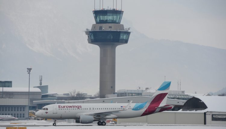 Ein Eurowings-Flugzeug rollt auf einer verschneiten Landebahn mit einem Flughafenkontrollturm im Hintergrund.