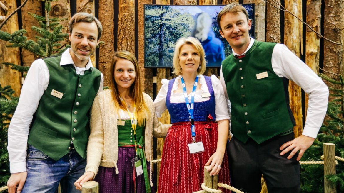 Vier Personen in traditioneller österreichischer Tracht stehen lächelnd vor einer rustikalen Holzkulisse zusammen.