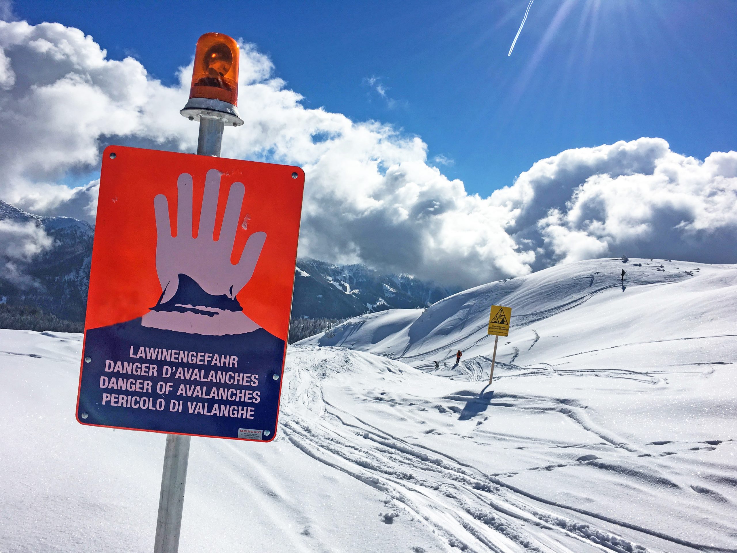 Warnschild mit roter Hand und Lawinengefahr auf verschneitem Berg bei teilweise bewölktem Himmel.