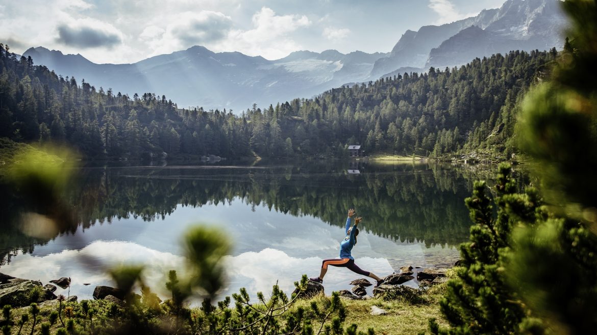 Person beim Yoga an einem ruhigen See, umgeben von Wald und Bergen unter einem teilweise bewölkten Himmel.