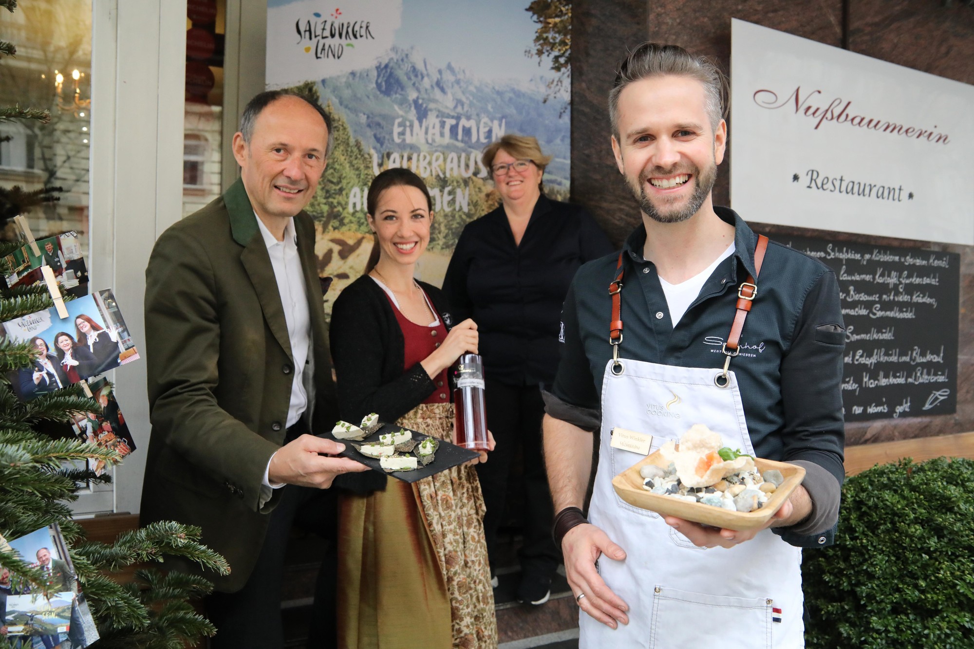 Vier Personen, die lächelnd Käseproben vor einem Restaurant mit alpenländischem Dekor servieren.