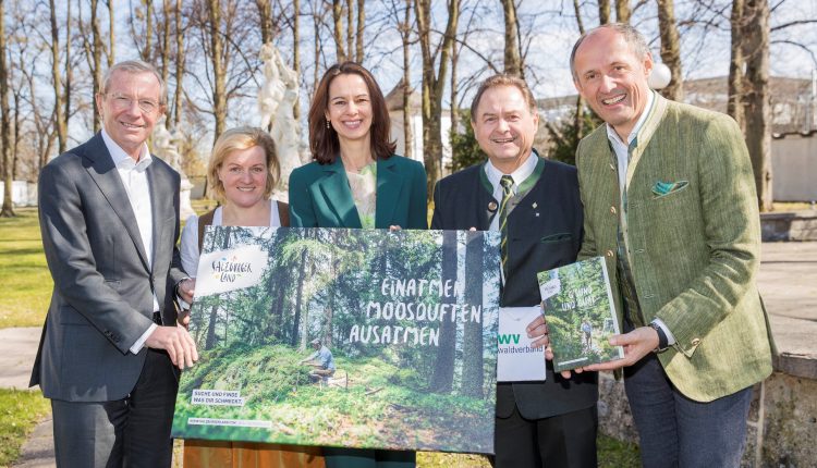 Fünf Personen im Freien, die lächelnd ein großes Poster mit einer Waldszene und einem Buch in der Hand halten, mit Bäumen im Hintergrund.