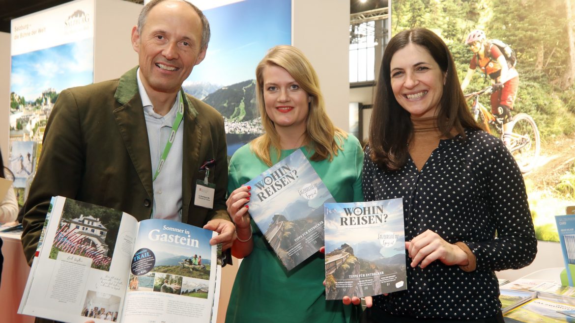 Drei Personen halten an einem Stand auf einer Tourismusmesse Reisemagazine und Broschüren hoch, im Hintergrund sind Bilder von Bergen zu sehen.