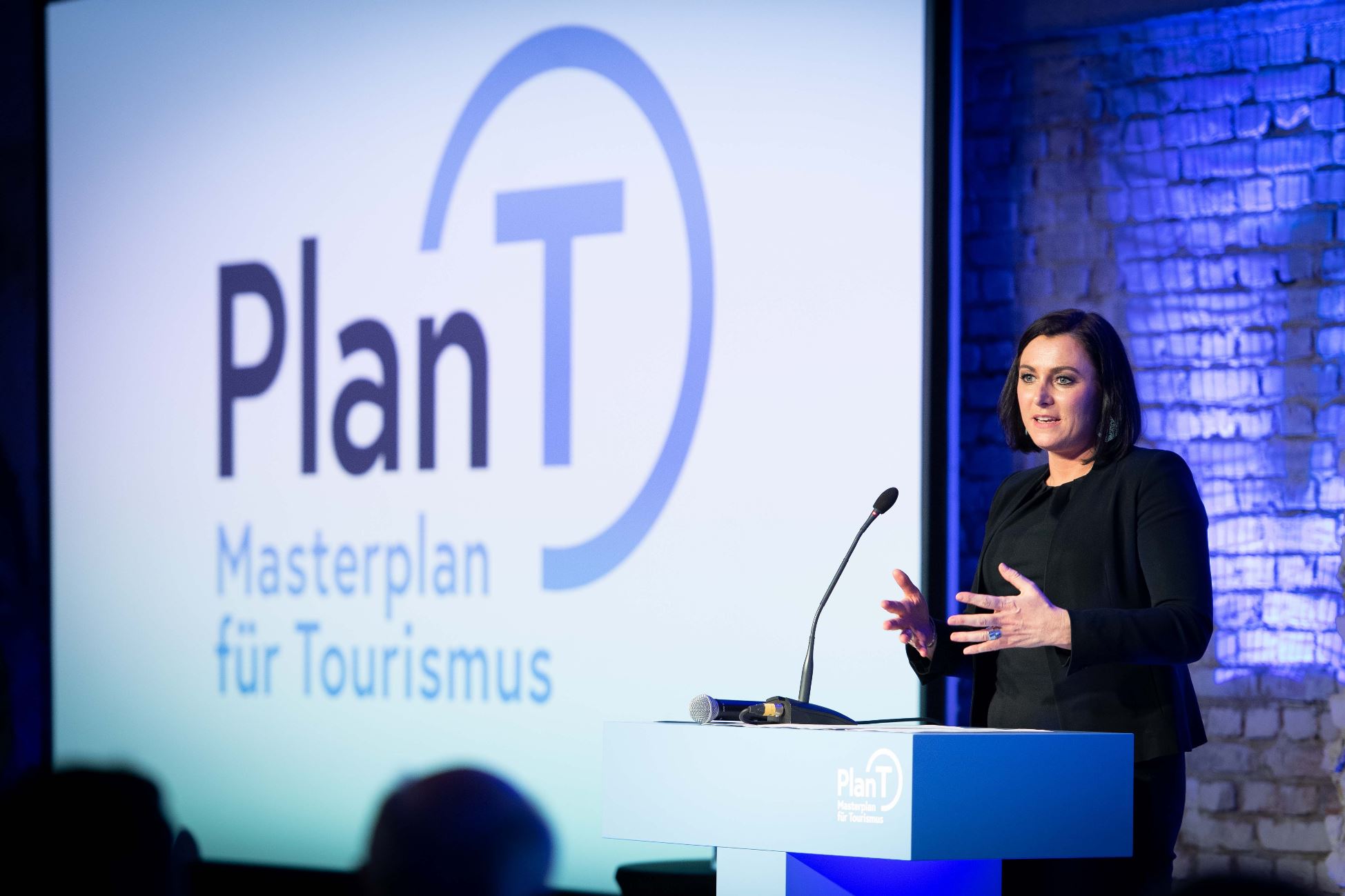 Eine Frau spricht an einem Podium mit einem Plan T Masterplan für Tourismus Schild und Logo im Hintergrund.