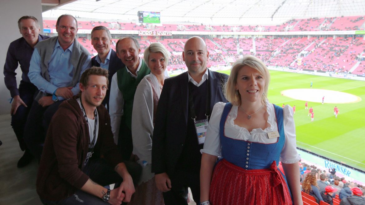 Eine Gruppe lächelnder Menschen in einer Stadionsuite, im Hintergrund ist ein Fußballfeld zu sehen.